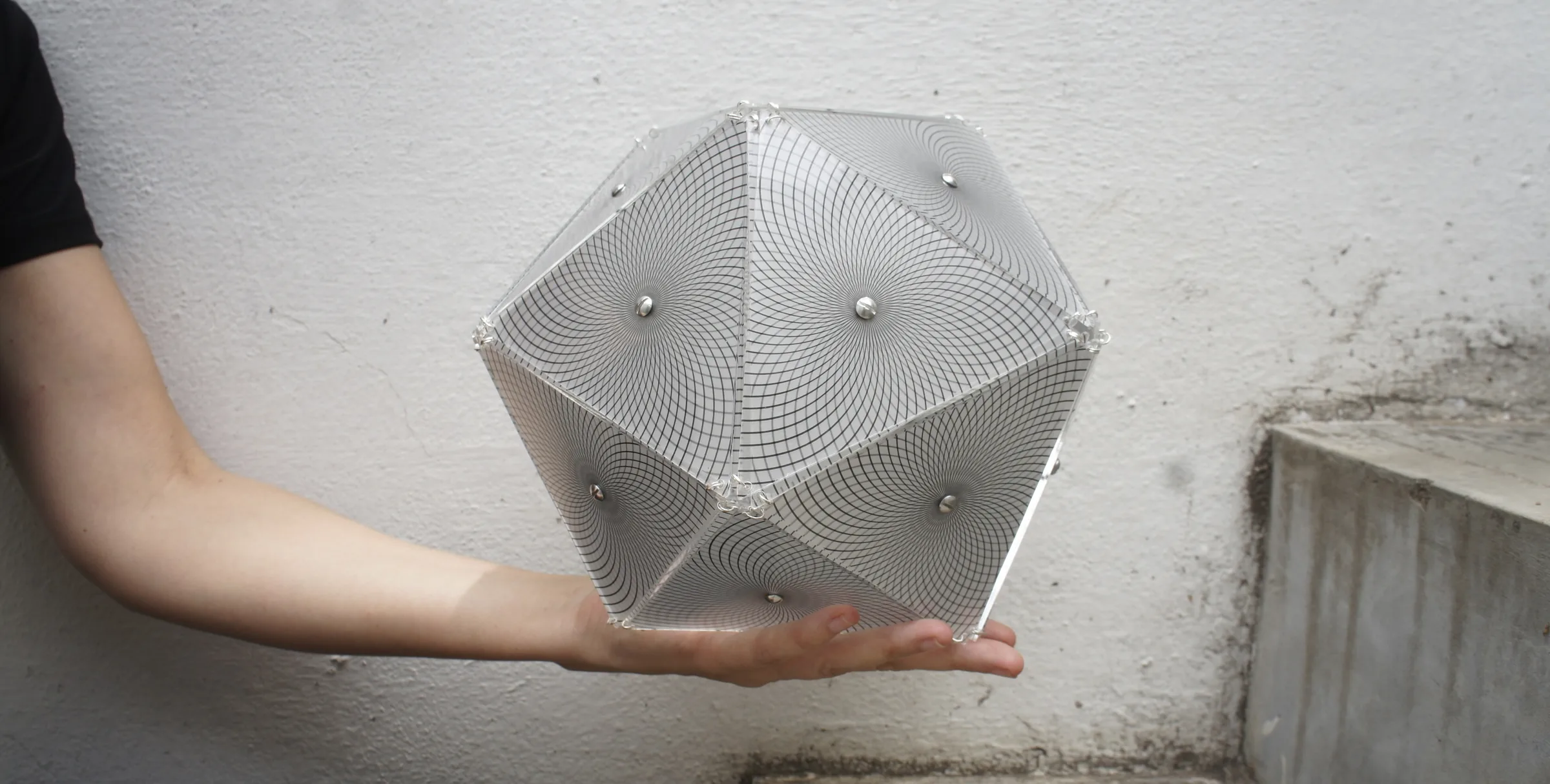 A handheld translucent icosahedron etched with spiral moir&eacute; lines, held against a pale concrete wall