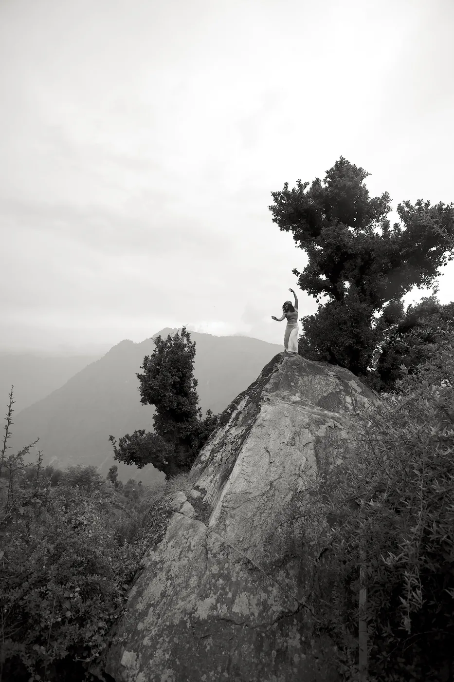 In black and white, a figure poised in a dance gesture atop a large boulder, a tree beside her and misty ridgelines beyond.