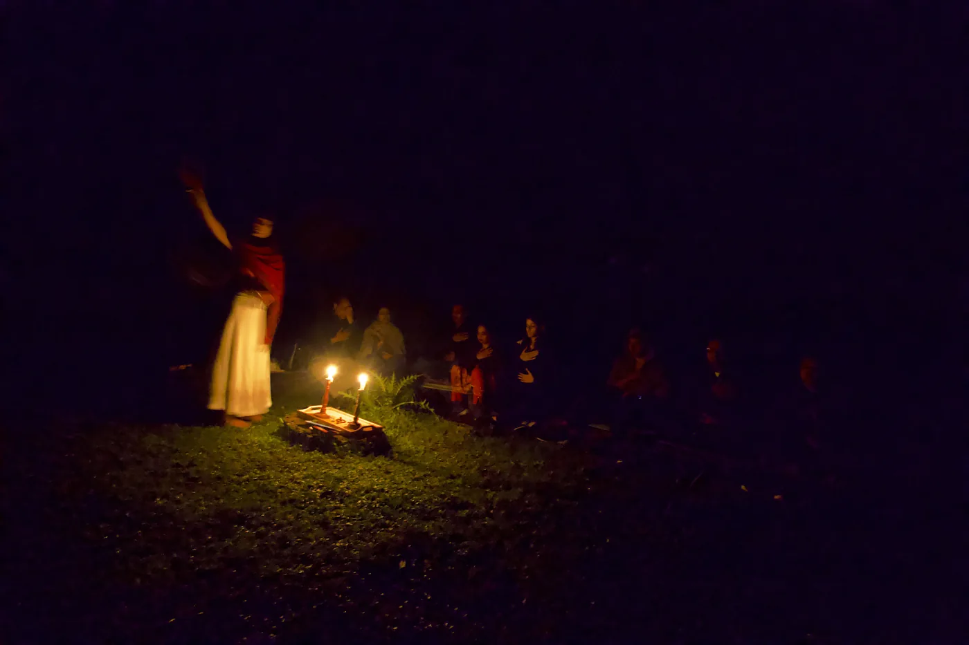 By night, a robed figure with one arm raised leads a small candlelit gathering seated on the grass.