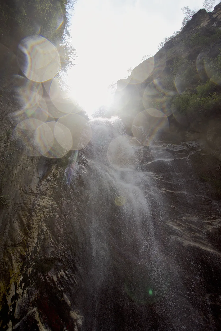 A waterfall falling through a steep rocky gorge into bright sun, scattered with rainbow lens-flare bokeh.