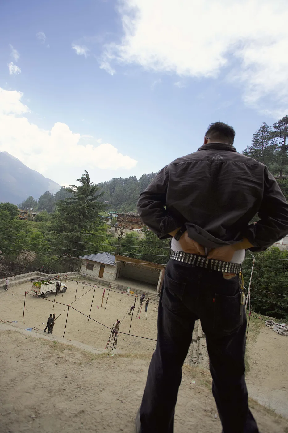 A man seen from behind, hands on hips, looking down over a sandy village ground being strung with lines, cedar forest and peaks beyond.