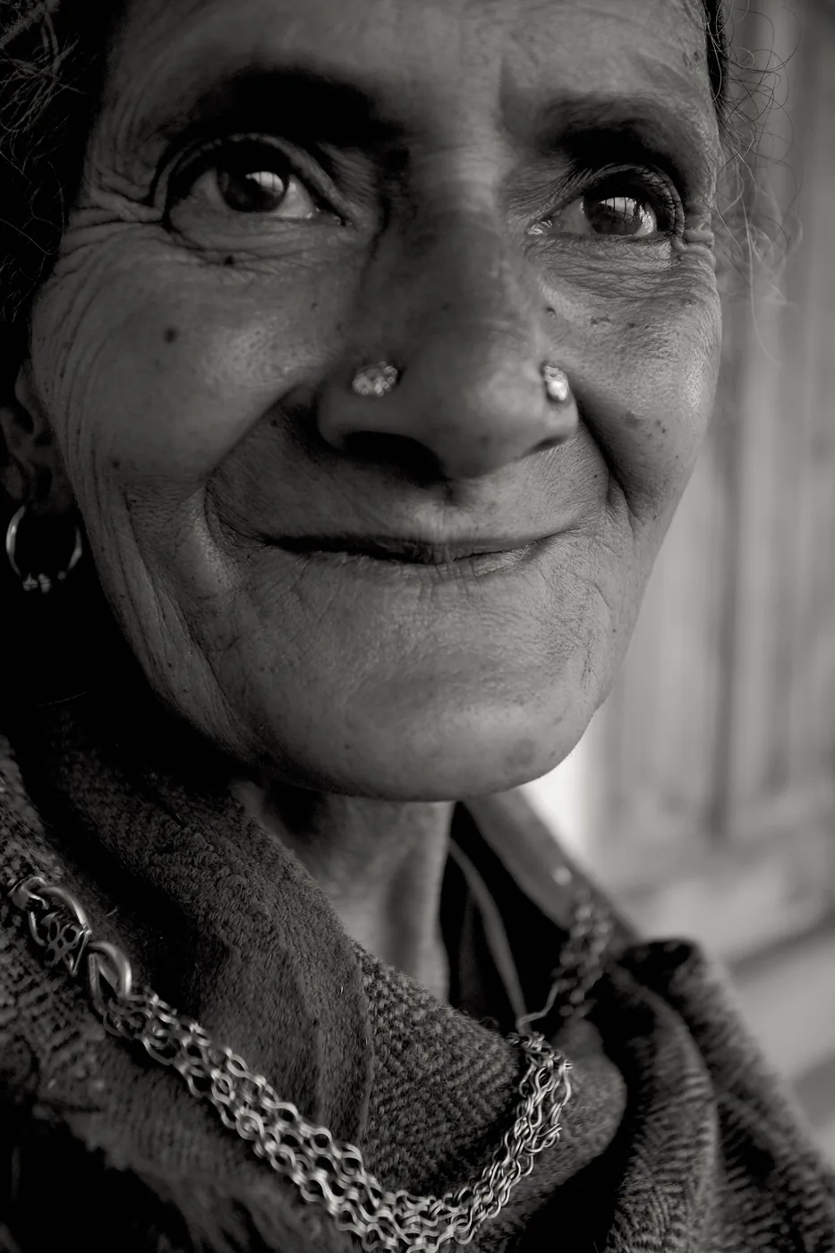 A black-and-white close portrait of an elderly woman smiling, with nose studs, a hoop earring and silver chains.