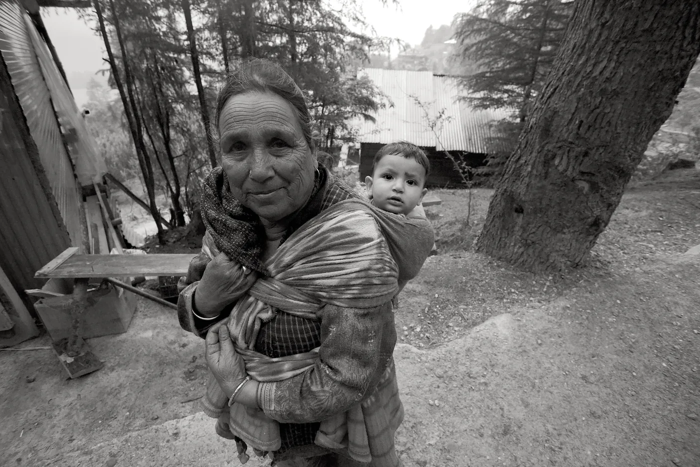 A black-and-white portrait of an elderly woman carrying a small child on her back in a wrap, cedars and a tin-roofed house behind.