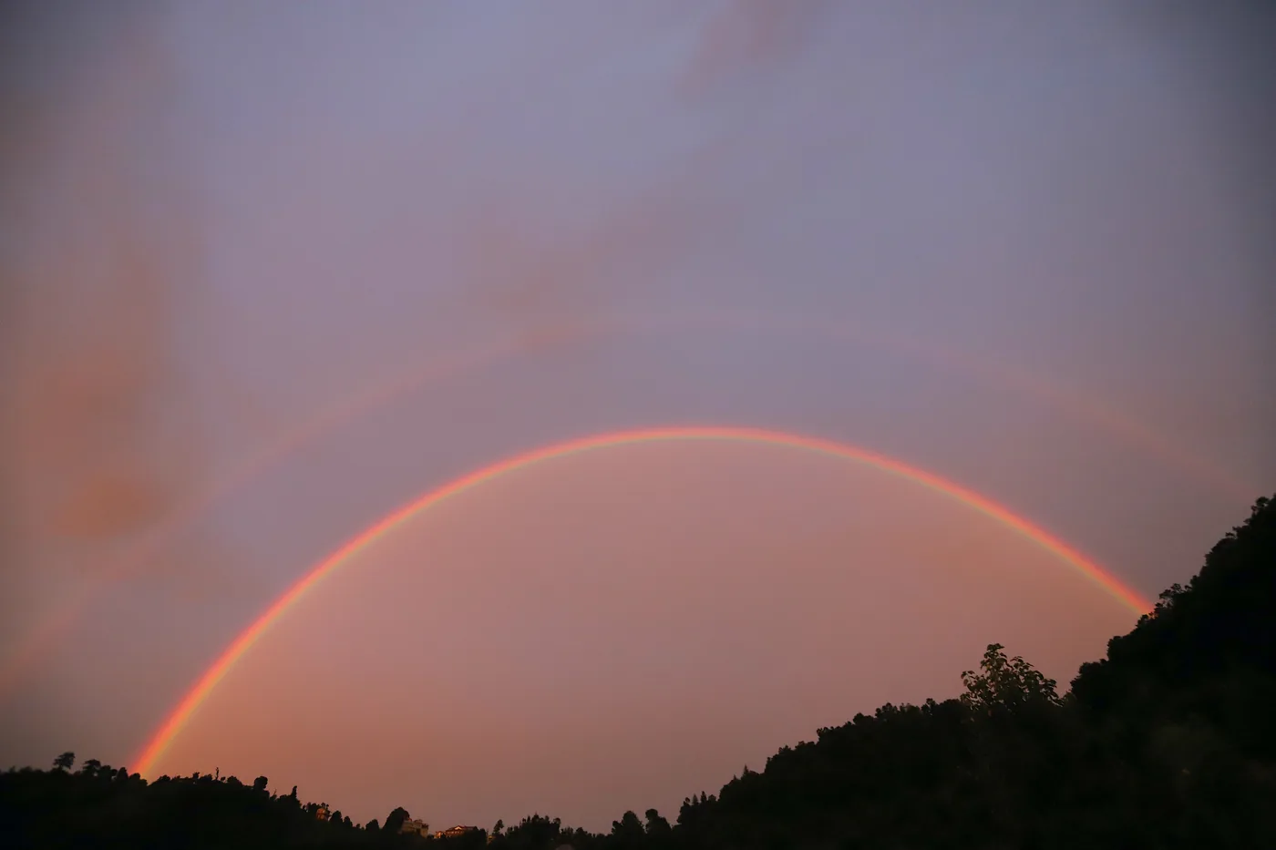 A rainbow arcing over forested hillsides under a soft violet dusk.