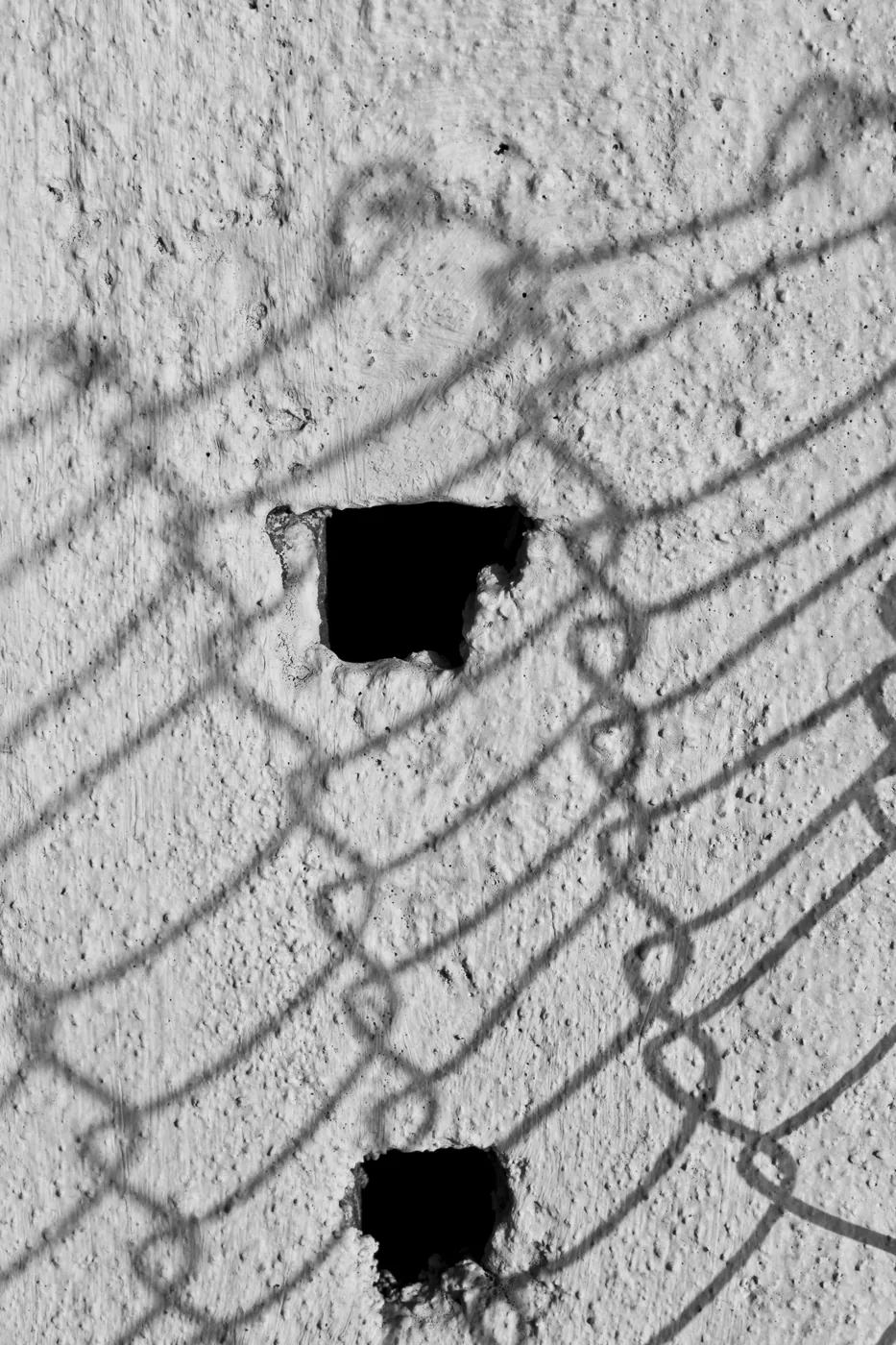 In black and white, a rough concrete wall marked by two dark holes and the shadow of a chain-link fence.