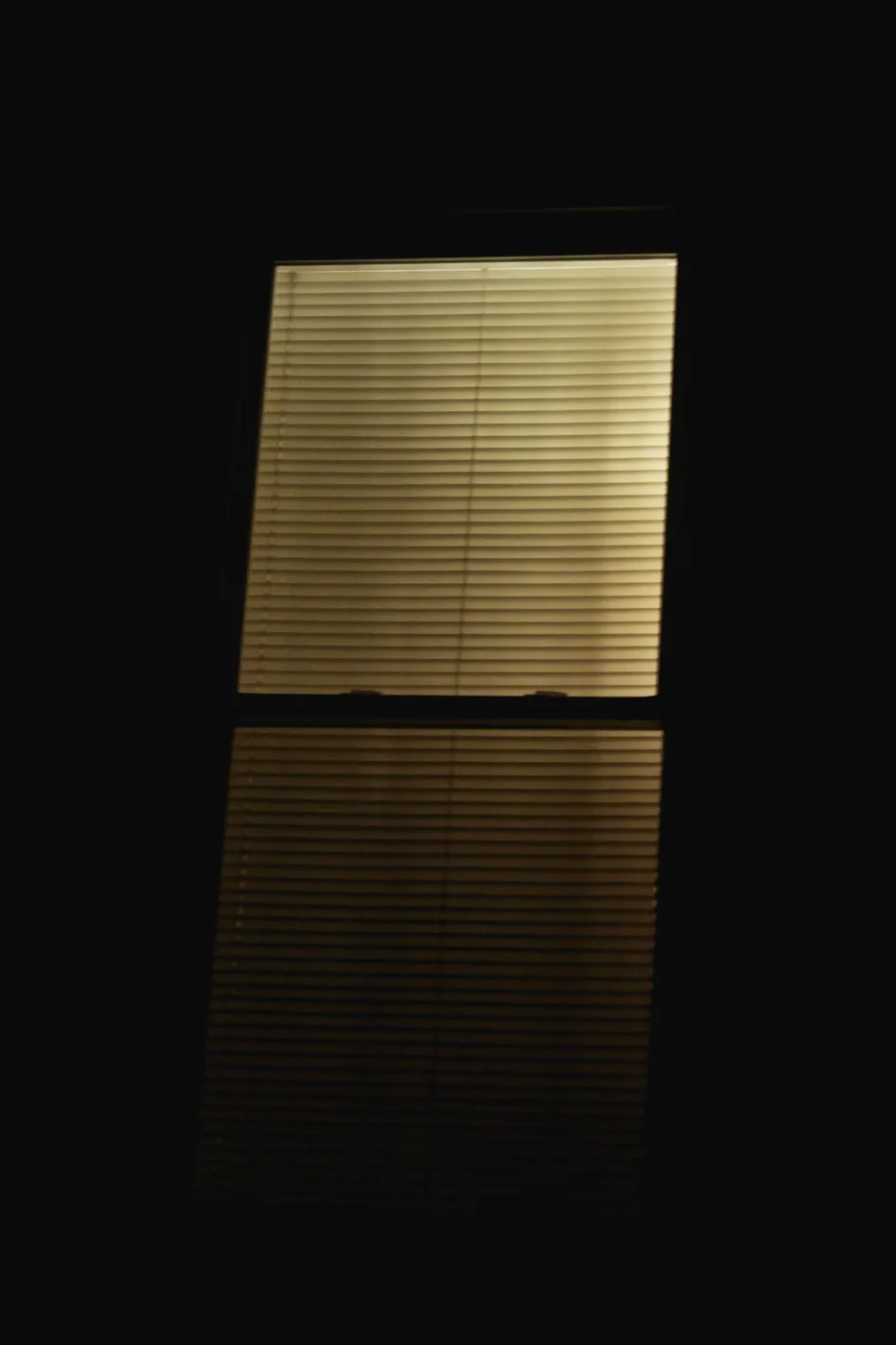 A glowing window blind in a dark room, mirrored in the surface below.