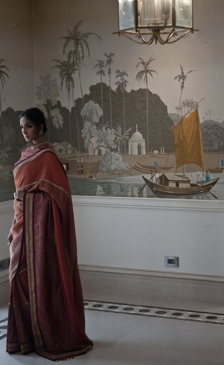 A woman in a red and maroon sari standing in a grand room before a painted mural of palms, a river and a boat with a yellow sail, a lantern hanging above.