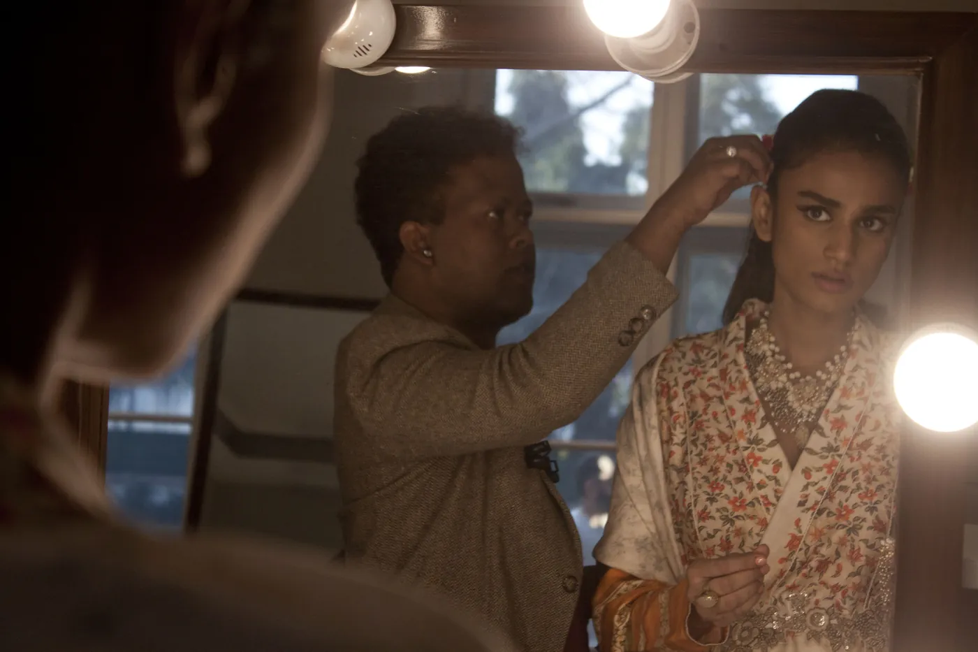 Backstage, reflected in a bulb-rimmed mirror, a stylist pinning the hair of a model in a floral robe.