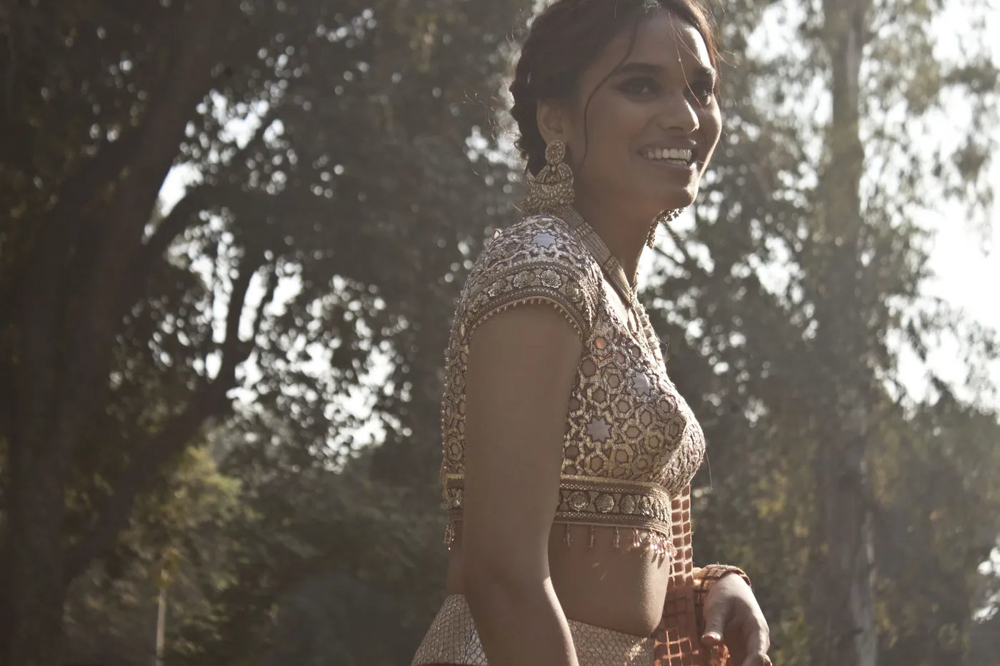 A woman in a gold mirror-worked blouse smiling in profile, backlit by sun filtering through tall trees.