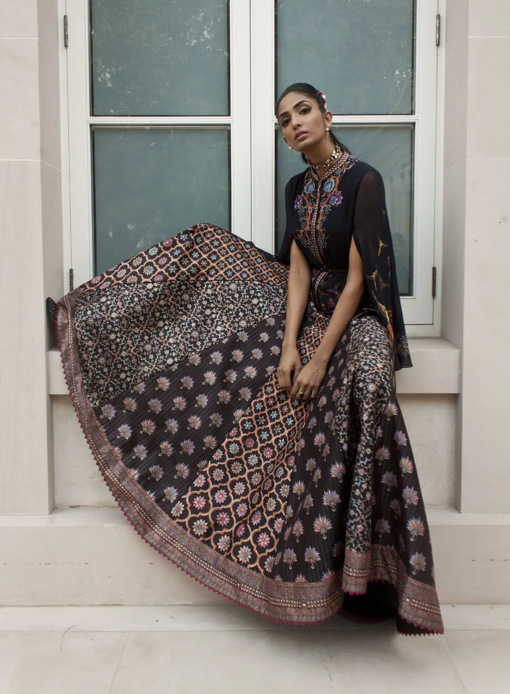 A woman seated on a windowsill, the wide skirt of her black floral-print lehenga spread across the stone ledge.