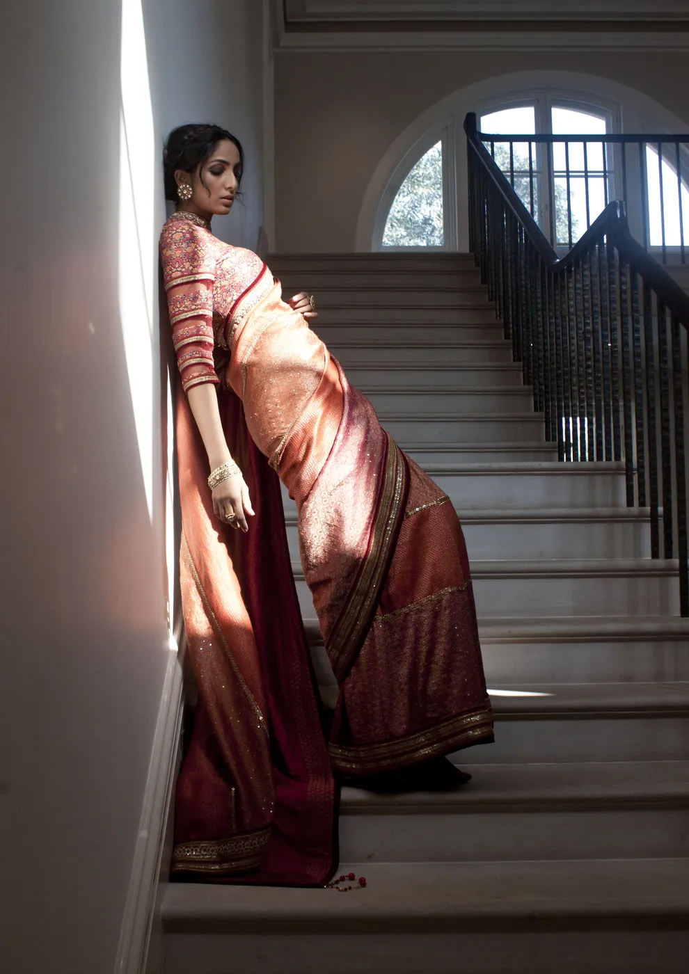 A woman in an orange and maroon silk sari leaning against the wall of a pale staircase, a shaft of daylight falling across her.