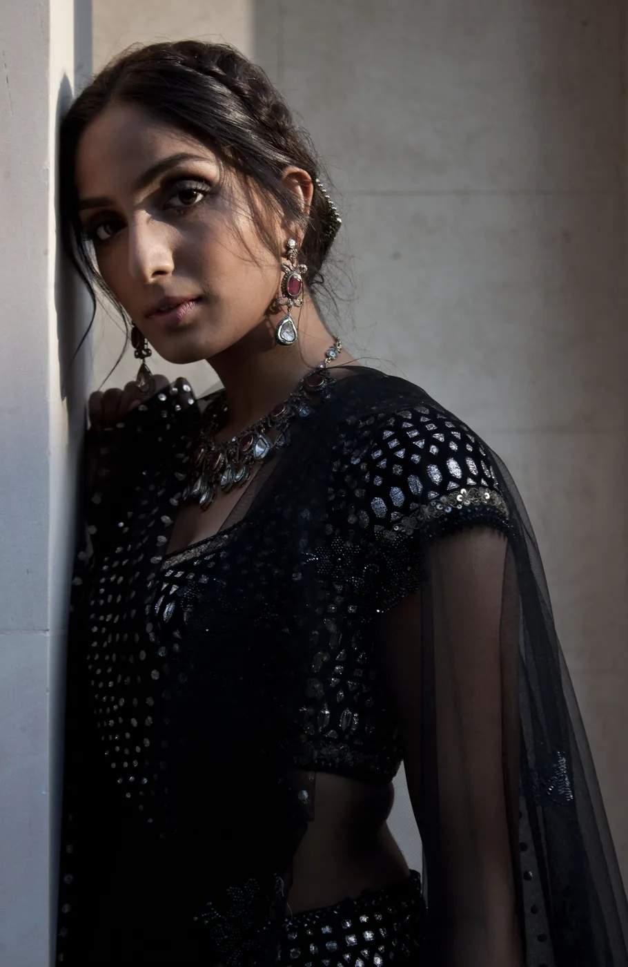 A close portrait of a woman in a black mirror-worked outfit with ruby and diamond earrings, leaning against a pale wall.