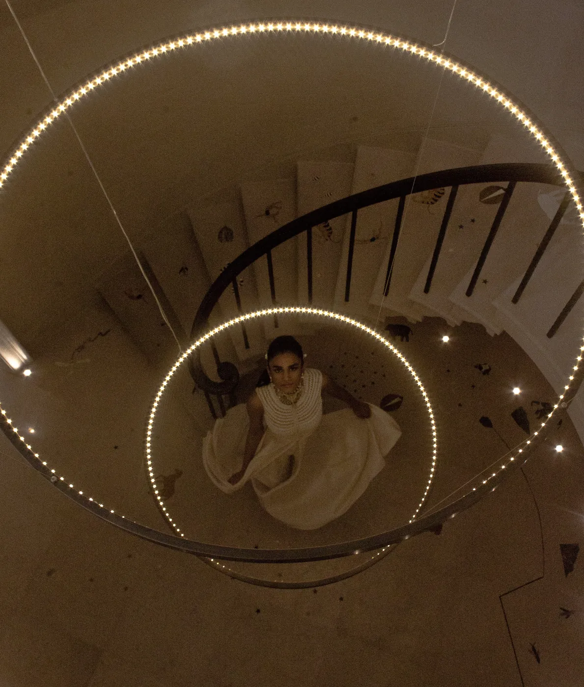 Looking down a spiral staircase ringed with strings of light, a woman in a flowing white gown standing at its centre far below.