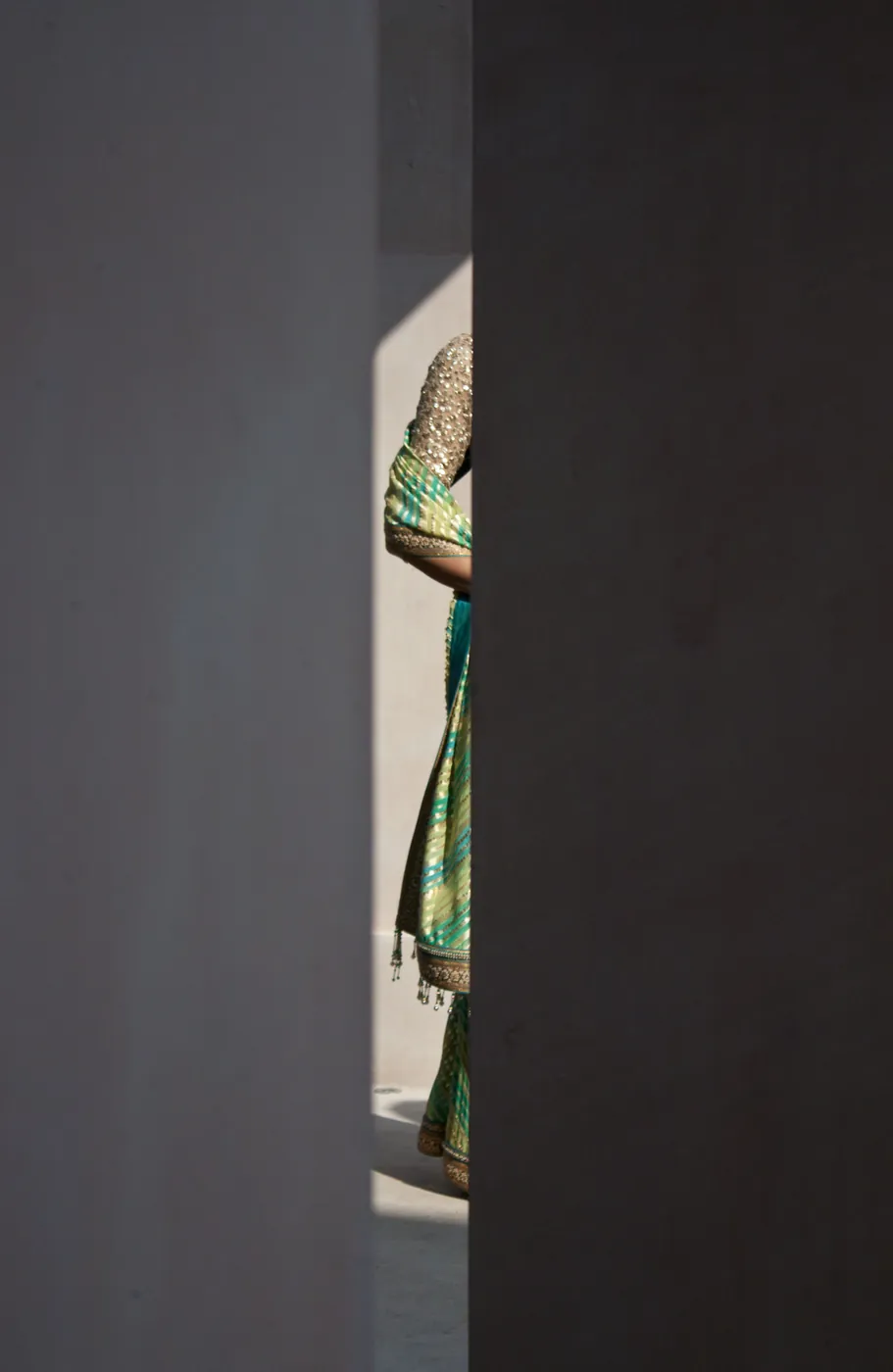 A woman in a green and gold sari glimpsed through a narrow gap between two walls, sunlight catching her sequinned veil.