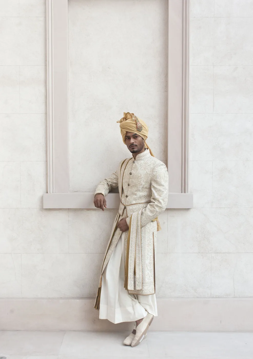 A man in an ivory embroidered sherwani and gold turban leaning beside a recessed panel in a pale stone wall.
