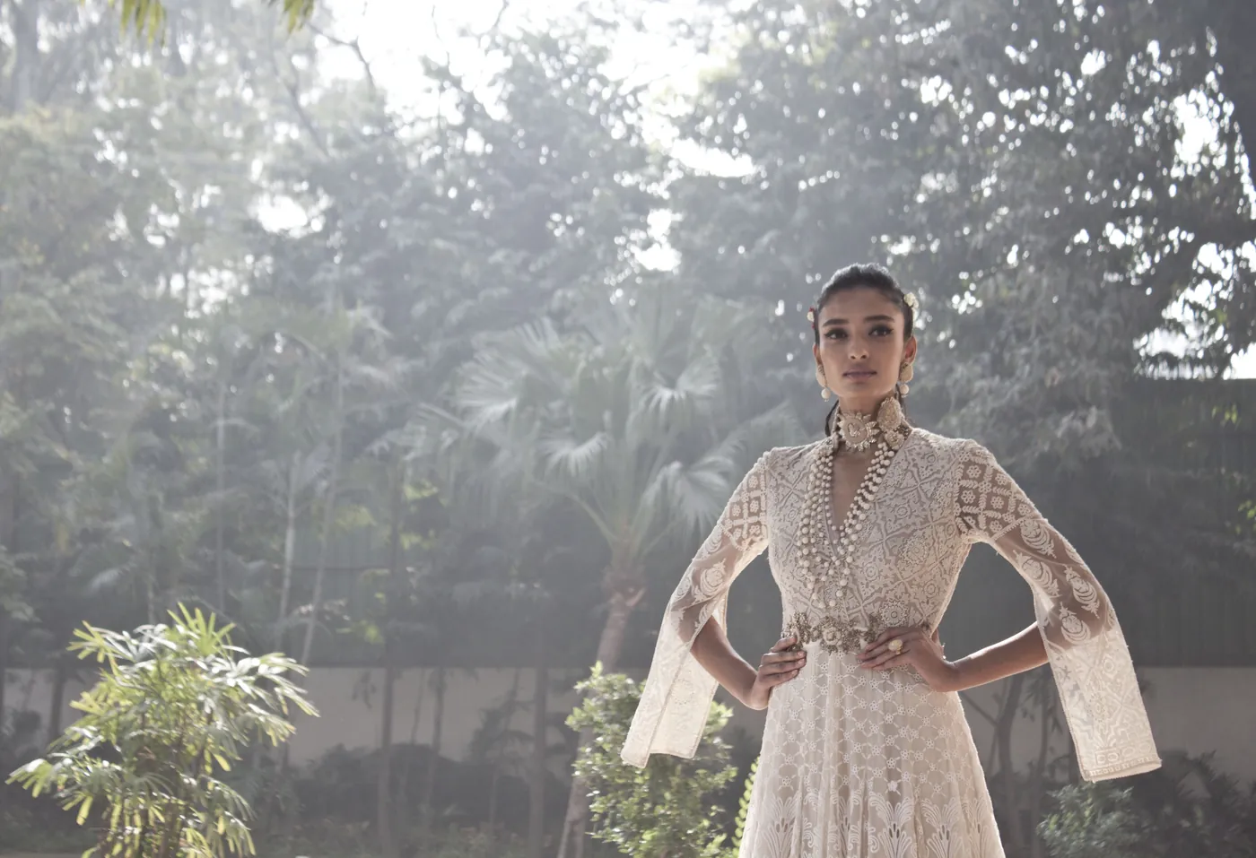 A woman in an ivory lace gown, hands at her hips, standing against a misty garden of palms and tall trees.