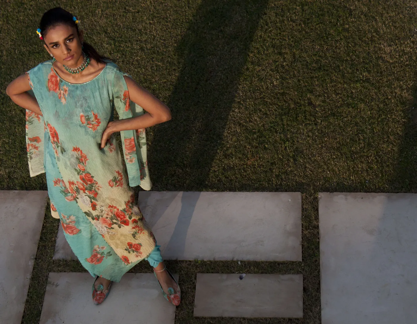 Seen from above, a woman in a turquoise floral suit standing where grass meets stone paving, her dupatta trailing.