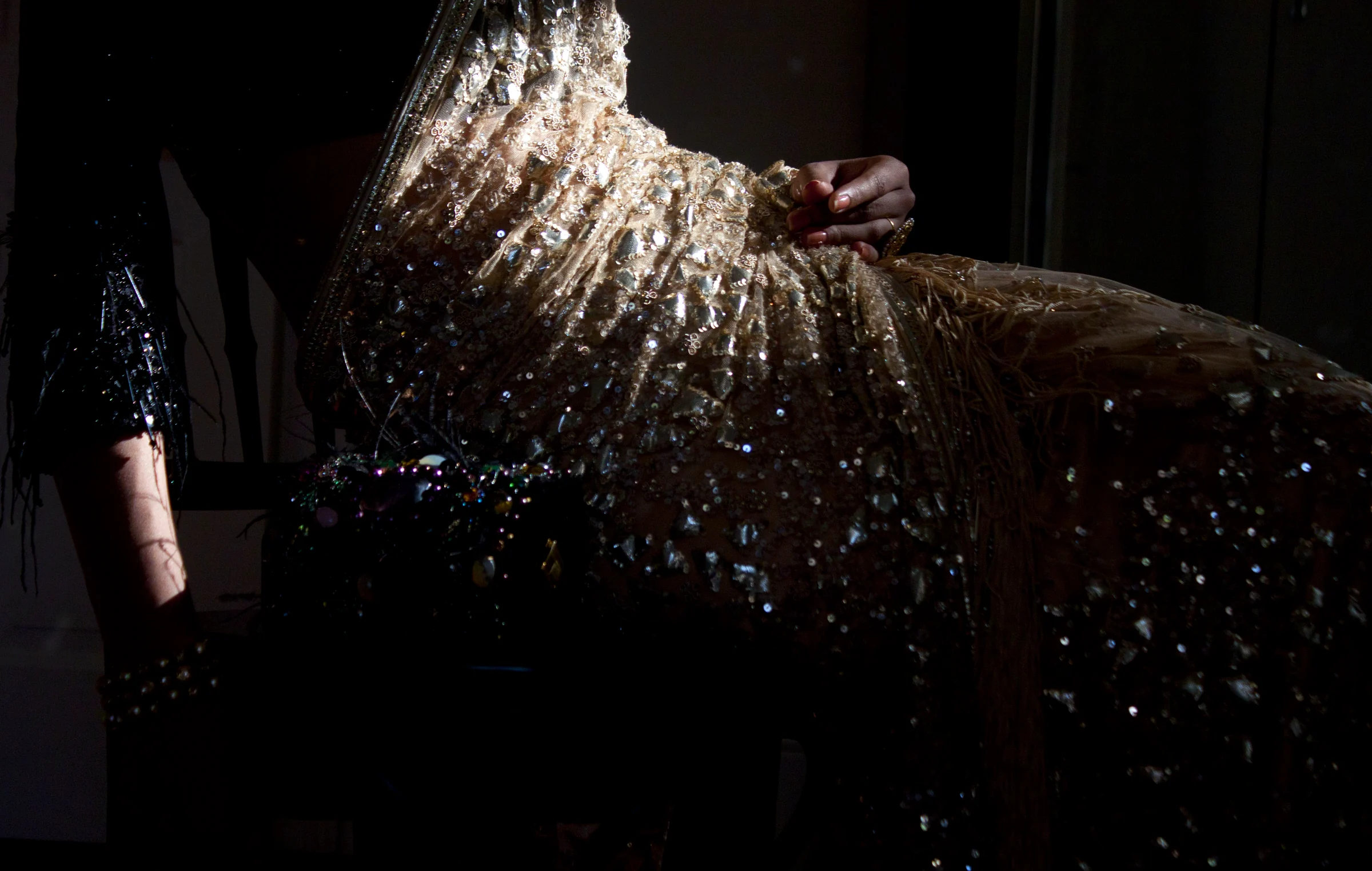 A hand gathering the folds of an intricately beaded, sequined garment that catches the light against a dark ground.