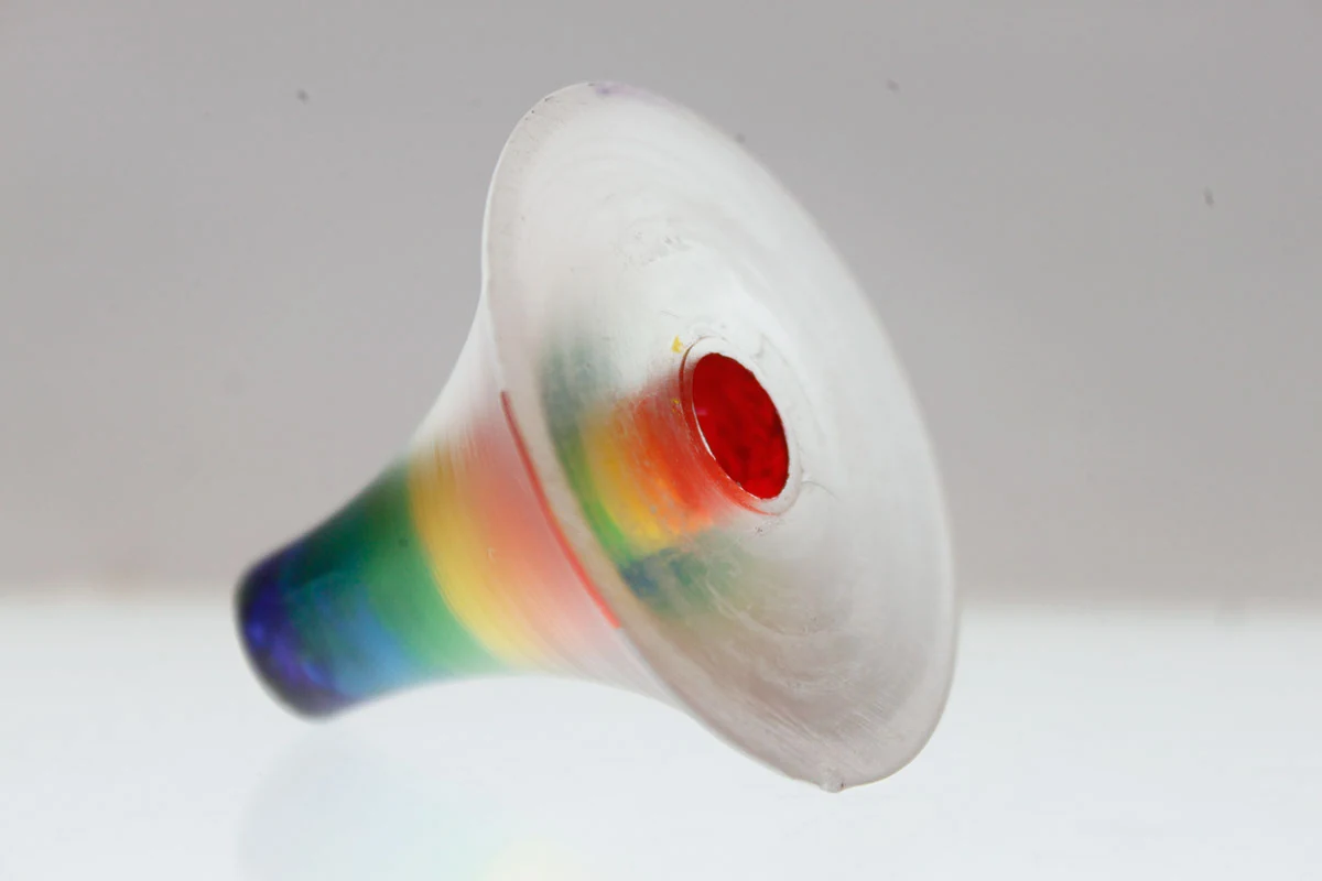 Photograph of the sculpture: a small translucent acrylic cone resting on a white ground, its flare wrapped in spectral bands from a violet-blue throat to a red mouth, with a disc of red set into the centre of its face.